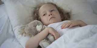 伤心的小女孩带着玩具在床上哭泣，不开心的生病的孩子感到孤独
