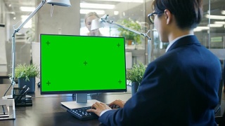 在办公室里，商人在他的办公桌上用一台模拟的绿色屏幕的个人电脑工作。同事走进办公室，并在她的办公桌前发生。视频素材模板下载