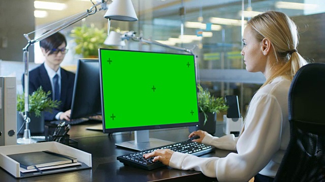 在办公室里，女商人在她的办公桌前对着一台模拟的绿色屏幕的个人电脑工作。同事走进办公室，并在他的办公桌前发生。