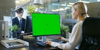 在办公室里，女商人在她的办公桌前对着一台模拟的绿色屏幕的个人电脑工作。同事走进办公室，并在他的办公桌前发生。