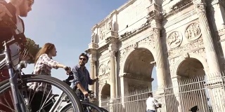在阳光明媚的一天，三个年轻的朋友骑着自行车在罗马市中心的罗马圆形大剧场康斯坦丁拱门附近散步