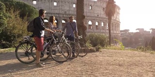 三个快乐的年轻朋友游客与自行车和背包在罗马斗牛场挥手，在山上的日落与树木的慢镜头娱乐