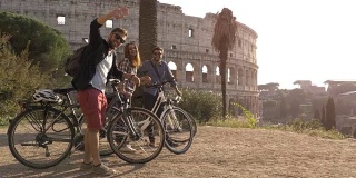 三个快乐的年轻朋友游客与自行车和背包在罗马斗牛场挥手，在山上的日落与树木的慢镜头娱乐