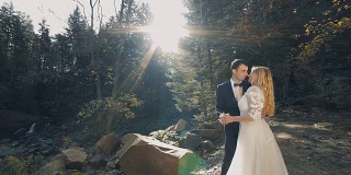 一对年轻美丽的新婚夫妇站在瀑布附近的森林里接吻。阳光灿烂的婚礼。慢动作