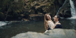一对美丽的年轻夫妇站在瀑布旁边。新郎亲吻新娘的脖子。结婚的那一天。慢动作