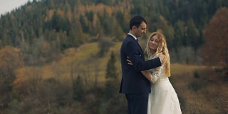 一对年轻美丽的新婚夫妇共同站在秋天的森林前。新娘倒在新郎的胸前。新郎拥抱她。缓慢的运动。结婚的那一天