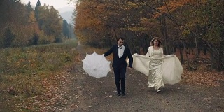 一对年轻美丽的新婚夫妇在秋天的森林里一起奔跑。新郎手里拿着一把白色的伞。结婚的那一天。慢动作