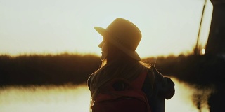 戴着帽子的快乐可爱女孩坐在夕阳下的河边码头上。金色的小时。享受美妙的旅行时刻。4K后视图特写