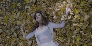 迷人的年轻女子穿着白色的连衣裙，微笑和放松躺在地上的秋天的树叶慢动作