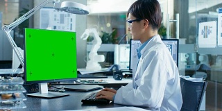 坐在办公桌前的亚洲科学家在一台模拟绿色屏幕的个人电脑上工作。在计算机科学研究实验室的机械臂模型。