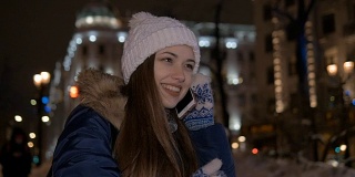 缓慢的运动。美丽的女孩微笑着和电话交谈。在冬天的大街上，在晚上。