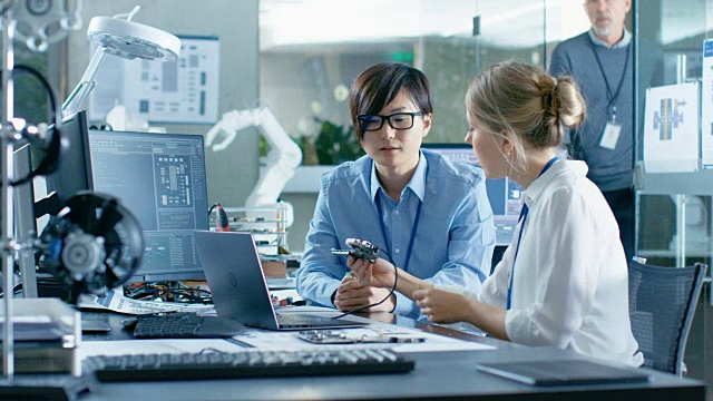 亚洲科学家坐在办公桌前向高级工程师咨询，他们将印刷电路板连接到笔记本电脑上并开始编码。在计算机科学研究实验室的机械臂模型。
