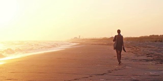 一个女人拿着一个包和沙滩鞋走在海滩在惊人的日落在夏季慢镜头。1920 x1080