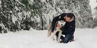 一名男子在森林里拥抱他的西伯利亚哈士奇。在雪的背景树。眼里是骄傲和幸福。带狗散步