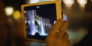 游客用便携式设备拍摄视频，音乐喷泉画面出现在屏幕上