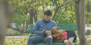 一对快乐的小情侣在公园的长椅上放松，女孩倚在男友的大腿上看书。