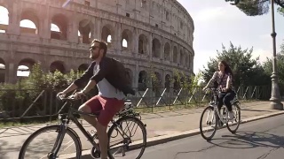 三个年轻的朋友游客在阳光明媚的日子里骑着自行车在罗马市中心的罗马斗牛场周围的道路上慢镜头摄影机汽车稳定摄影机视频素材模板下载