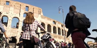 在阳光明媚的日子里，三个年轻的朋友带着自行车和背包在罗马斗牛场散步
