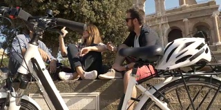 三个年轻的朋友，游客与自行车和背包坐在罗马斗牛场在阳光明媚的日子享受慢镜头摄影机稳定摄影机