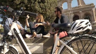 三个年轻的朋友，游客与自行车和背包坐在罗马斗牛场在阳光明媚的日子享受慢镜头摄影机稳定摄影机视频素材模板下载