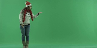 迷人的年轻女子在圣诞老人的帽子是站着，并告诉一些东西，而指向它在绿色屏幕上，色度键的背景