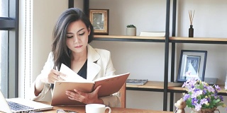 4K视频，美丽的亚洲商务女性坐在电脑笔记本电脑阅读和翻页笔记本和微笑在咖啡馆咖啡厅在城市的早晨，商务人士的生活方式。