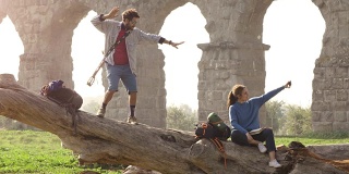 快乐的年轻一对背包客游客在一个原木树干上用智能手机自拍在古罗马渡槽遗址在浪漫的parco degli acququedotti公园在朦胧的日出慢镜头TRIPOD