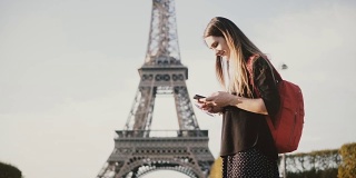 年轻漂亮的女子背着双肩包站在埃菲尔铁塔附近，用智能手机上网