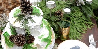 这是一个两层楼的白色婚礼蛋糕的特写镜头，上面装饰着松果和云杉树枝，上面的桌子上摆满了用蜡烛装饰的盘子和玻璃杯。