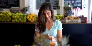美丽的女人在一个果汁吧享受果汁，同时用她的智能手机聊天微笑