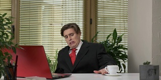 一个穿着西装的年轻人正坐在办公室里，用笔记本电脑工作，不是喝着美味的咖啡，做着鬼脸。在办公室工作。60 fps