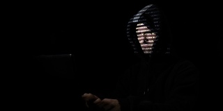 戴着兜帽的男性网络罪犯坐在一间黑屋子里攻击区块链的服务器而代码字符会反射到他的脸上
