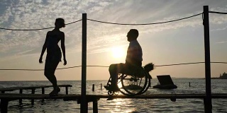 残疾人士过着充实的生活年轻妇女和男性残疾在轮椅上码头在背景天堂在橙色的阳光