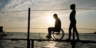 瘦弱的女孩和坐轮椅的残疾人在夕阳下的海上码头上行走
