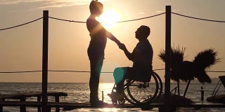 女性和男性残疾人的剪影在轮椅上抱着胳膊，在海上看地平线