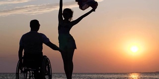长腿女人站在防波堤上，挥舞着一块布，在一个跛脚到轮椅上的男人面前，背景是河上橙色的日落
