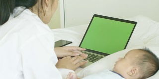 年轻的亚洲母亲和她刚出生的孩子在卧室的绿屏笔记本电脑上工作。