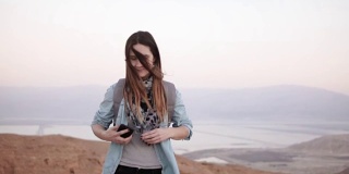 黄昏时分，一名女子在使用智能手机欣赏山景。缓慢的运动。messenger应用中背包类型的休闲女孩。以色列