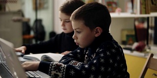 两个小男孩穿着温暖舒适的毛衣坐在编程课上的笔记本电脑前，试图学习如何使用这些小工具的慢镜头。
