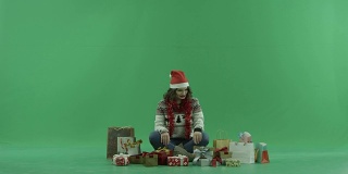 迷人的年轻女子在圣诞帽坐在圣诞礼物和试图选择一个，色度键的背景