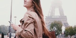 年轻漂亮的女人在艾菲尔铁塔附近使用智能手机。微笑的女性环顾四周，看着法国的巴黎