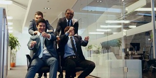 四名滑稽的多民族商人坐在公司大楼走廊的椅子上。同事们欢呼鼓掌。庆祝交易完成。