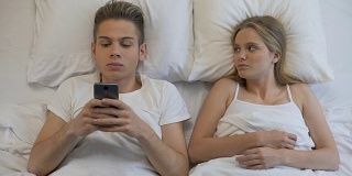 愤怒的女孩看着男友在床上用手机，网瘾