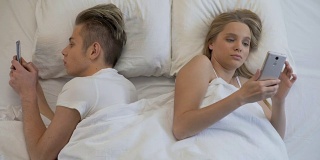 一对年轻夫妇背靠背躺在床上用手机上网，漠不关心