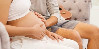美丽的孕妇和她的丈夫抱着肚子在家里的沙发上听着宝宝，怀孕和家庭的概念