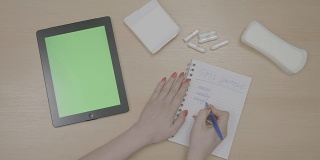 用绿色屏幕的平板电脑和在记事本上写，而桌子上有卫生棉条的女人的红色指甲的手计划经前综合症PMS名单