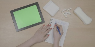 俯视图，一个年轻女人的手计划周期跟踪清单与绿色屏幕平板电脑和书写在记事本周围的卫生棉条