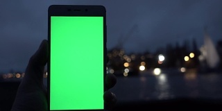 男子手握白色智能手机，在夜城灯光散景的背景下。绿色的屏幕。