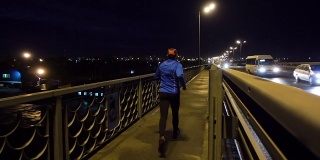 晚上在桥边慢跑的运动员。一个穿着运动服的男人在晚上跑步。男正在健身，车路过。
