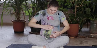 一位穿着灰色运动衬衫的漂亮孕妇站在窗前，抚摸着自己的肚子，看着镜头。未来的母亲，怀孕晚期。